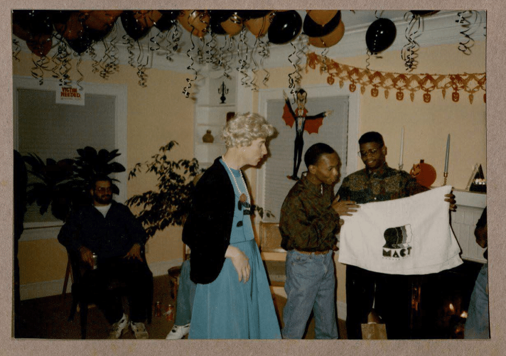 Photo from Men of All Colors Together-KC’s Halloween Party. On the right-hand side of the photo is Quience X. Sykes and Steve Ricard––both men admiring a white towel with MACT-KC’s logo printed over it ([Scrapbook Clippings of BWMT/MACT-KC], ca. 1990-1993)].
