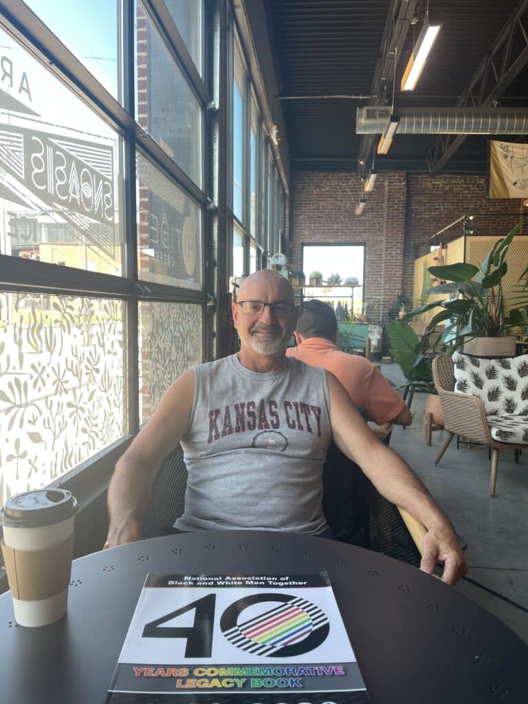 Reynolds smiles at the camera, sporting a Kansas City tank top and glasses. The table in front of him has a cup of coffee and NABWMT's '40 Years Commemorative Legacy Book,' a publication Reynolds says he worked on for three years.