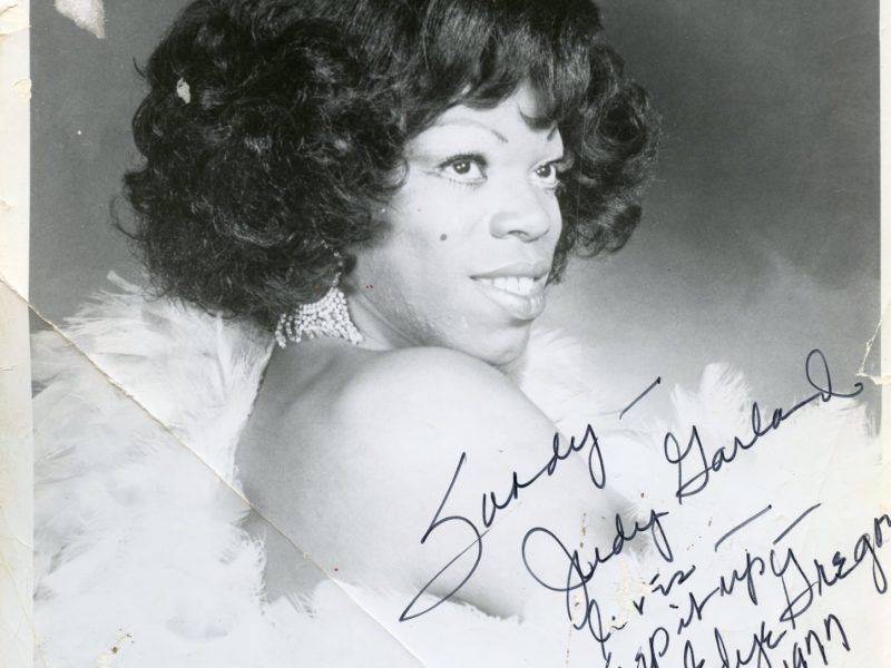 Edye Gregory in a signed headshot to a loved one. Edye smiles with her right shoulder facing the camera, dressed in a light-colored, feathered dress.