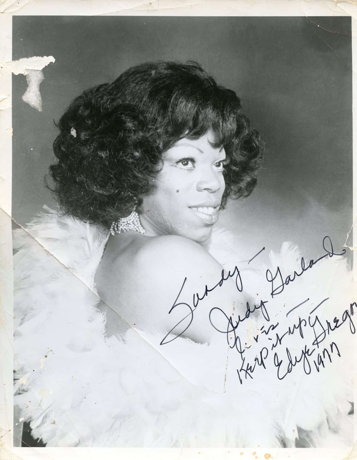 Edye Gregory in a signed headshot to a loved one. Edye smiles with her right shoulder facing the camera, dressed in a light-colored, feathered dress.