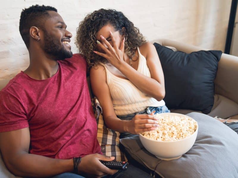 young couple watching horror movie while sitting on a couch at home. Young man is smiling with arm wrapped around a young girl covering her face with her hand.